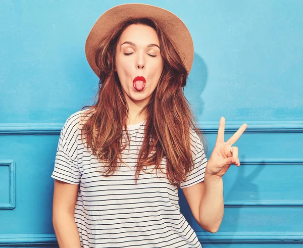 スタイリッシュな少女モデルではカジュアルな夏服と赤い唇 青い壁に近いポーズで茶色の帽子 ピースサインと彼女の舌を示す — ストック写真