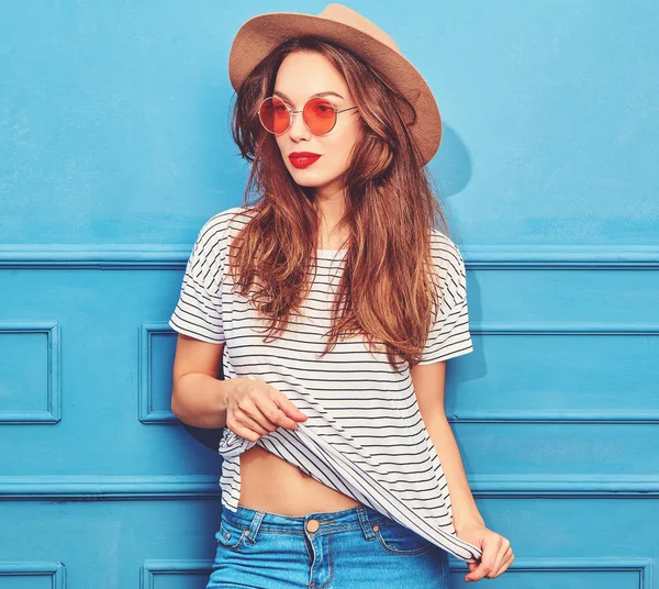 スタイリッシュな少女モデルのカジュアルな夏服と赤い唇 青い壁に近いポーズで茶色の帽子 — ストック写真