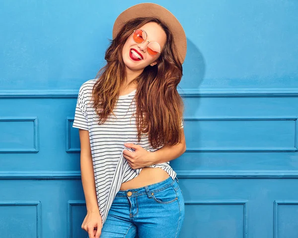 年轻时尚的女孩在休闲夏天的衣服和棕色的帽子 红色的嘴唇 在蓝色的墙壁上的造型 显示她的舌头 — 图库照片