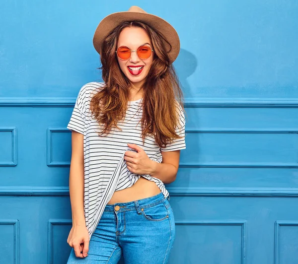 スタイリッシュな少女モデルではカジュアルな夏服と赤い唇 青い壁に近いポーズで茶色の帽子 彼女の舌を見せて ウインク — ストック写真