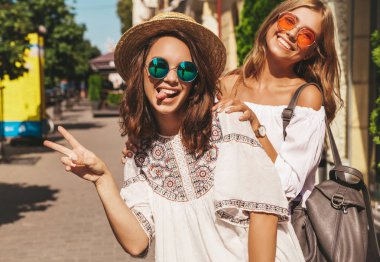 İki genç şık hippi esmer ve sarışın Bayan modelleri yaz güneşli gün sokak arka plan üzerinde poz beyaz hipster giysiler içinde. Barış işareti ve dilini gösteren