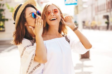 İki genç şık hippi esmer ve sarışın kadın sokak arka plan üzerinde poz beyaz hipster giyim yaz güneşli bir günde moda portre. Sırlar, dedikodu paylaşmak modelleri.