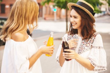 İki genç şık hippi esmer ve sarışın Bayan modelleri yaz güneşli gün sokak arka plan üzerinde poz beyaz hipster giysiler içinde. Şişe soda içme ve iletişim
