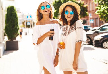 İki genç şık hippi esmer ve sarışın Bayan modelleri yaz güneşli günde sokak arka plan üzerinde poz beyaz hipster giysileri moda portre. Şişe soda içme