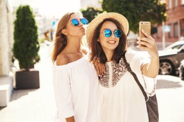 İki genç kadın gülümseyen hippi esmer ve sarışın kadın modeli selfie sosyal medya için Smartphone cep telefonu ile sokak arka plan üzerinde çekim beyaz hippi elbise yaz. Sürpriz yüz, duygular,
