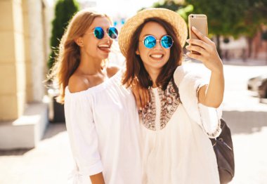 İki genç kadın şık hippi esmer ve sarışın Bayan modelleri yaz güneşli günde selfie sosyal medya için Smartphone cep telefonu ile sokak arka plan üzerinde çekim beyaz hipster giysili