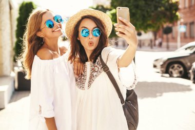 İki genç kadın gülümseyen hippi esmer ve sarışın kadın modeli selfie sosyal medya için Smartphone cep telefonu ile sokak arka plan üzerinde çekim beyaz hippi elbise yaz. Sürpriz yüz, duygular,