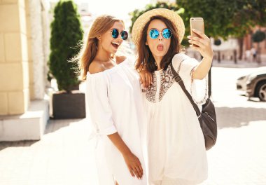 İki genç kadın gülümseyen hippi esmer ve sarışın kadın modeli selfie sosyal medya için Smartphone cep telefonu ile sokak arka plan üzerinde çekim beyaz hippi elbise yaz. Sürpriz yüz, duygular,