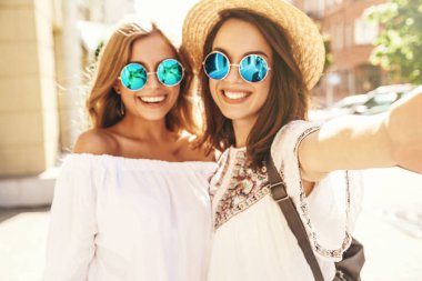 İki genç kadın gülümseyen hippi esmer ve sarışın kadın modeli selfie sosyal medya için Smartphone cep telefonu ile sokak arka plan üzerinde çekim beyaz hippi elbise yaz. Sürpriz yüz, duygular,