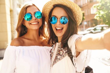 İki genç kadın gülümseyen hippi esmer ve sarışın Bayan modelleri yaz güneşli günde selfie sosyal medya için Smartphone cep telefonu ile sokak arka plan üzerinde çekim beyaz hipster giysili