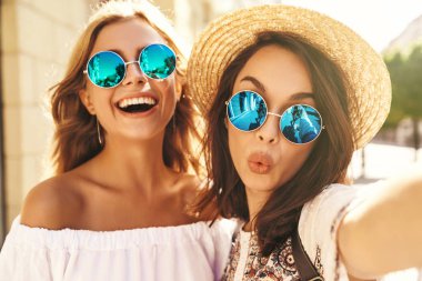 İki genç kadın gülümseyen hippi esmer ve sarışın Bayan modelleri yaz güneşli günde selfie sosyal medya için Smartphone cep telefonu ile sokak arka plan üzerinde çekim beyaz hipster giysili. Öpücük vererek