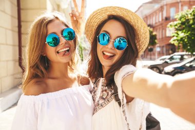 İki genç kadın şık hippi esmer ve sarışın Bayan modelleri yaz güneşli günde selfie sosyal medya için Smartphone cep telefonu ile sokak arka plan üzerinde çekim beyaz hipster giysili. Dil gösterilen