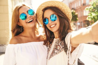 İki genç kadın gülümseyen hippi esmer ve sarışın kadın modeli selfie sosyal medya için Smartphone cep telefonu ile sokak arka plan üzerinde çekim beyaz hippi elbise yaz. Sürpriz yüz, duygular,