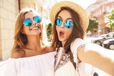 İki genç kadın gülümseyen hippi esmer ve sarışın kadın modeli selfie sosyal medya için Smartphone cep telefonu ile sokak arka plan üzerinde çekim beyaz hippi elbise yaz. Sürpriz yüz, duygular,