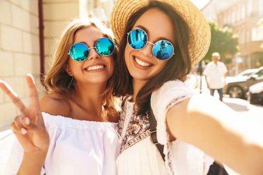 İki genç kadın gülümseyen hippi esmer ve sarışın Bayan modelleri yaz güneşli günde selfie sosyal medya için Smartphone cep telefonu ile sokak arka plan üzerinde çekim beyaz hipster giysili. Barış işareti gösterilen