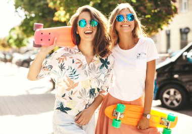 İki genç şık gülümseyen hippi esmer ve sarışın kadın modelleri yaz güneşli günde hipster giysili sokak arka plan üzerinde poz kuruş kaykay ile 
