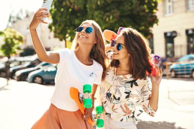 İki genç kadın şık hippi esmer ve sarışın Bayan modelleri yaz güneşli günde selfie sosyal medya için Smartphone cep telefonu ile sokak arka plan üzerinde çekim hipster giysili. Renkli kuruş kaykaylar ile 