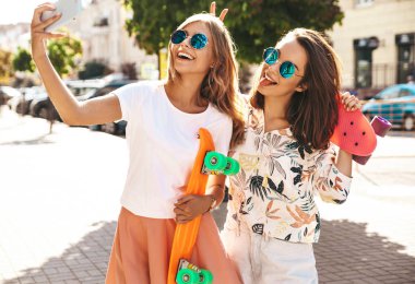 İki genç kadın şık hippi esmer ve sarışın Bayan modelleri yaz hipster giysileri selfie sosyal medya için Smartphone cep telefonu ile sokak arka plan üzerinde çekim. Renkli kuruş kaykaylar ile. Barış işareti göster