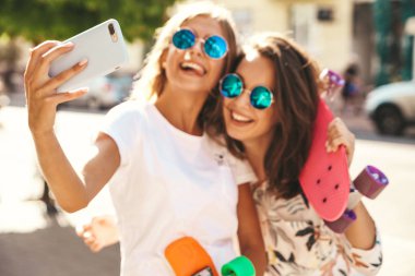 İki genç kadın şık hippi esmer ve sarışın Bayan modelleri yaz hipster giysileri selfie sosyal medya için Smartphone cep telefonu ile sokak arka plan üzerinde çekim. Renkli kuruş kaykaylar ile 