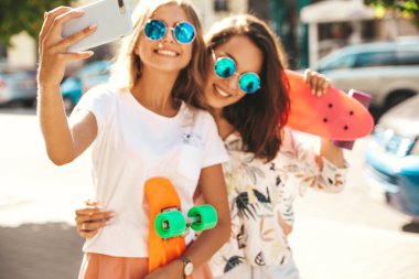 İki genç kadın şık hippi esmer ve sarışın Bayan modelleri yaz hipster giysileri selfie sosyal medya için Smartphone cep telefonu ile sokak arka plan üzerinde çekim. Renkli kuruş kaykaylar ile 
