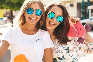 Yaz güneşli günde hipster giysili iki genç şık gülümseyen hippi esmer ve sarışın kadın modelleri sokak arka plan üzerinde poz kuruş kaykay ile. Dilleri gösterilen