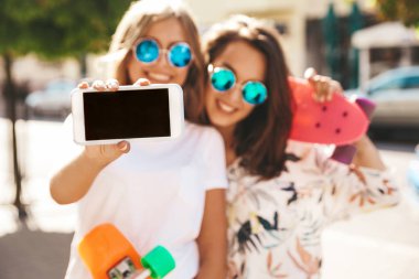 İki genç kadın şık hippi esmer ve sarışın Bayan modelleri yaz hipster giysileri selfie sosyal medya için Smartphone cep telefonu ile sokak arka plan üzerinde çekim. Renkli kuruş kaykaylar ile 