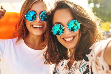 İki genç kadın gülümseyen hippi esmer ve sarışın Bayan modelleri yaz güneşli günde selfie sosyal medya için Smartphone cep telefonu ile sokak arka plan üzerinde çekim hipster giysili. Renkli kuruş kaykaylar ile 
