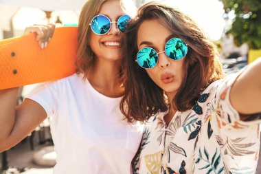 İki genç kadın şık hippi esmer ve sarışın Bayan modelleri yaz hipster giysileri selfie sosyal medya için Smartphone cep telefonu ile sokak arka plan üzerinde çekim. Renkli kuruş kaykaylar ile 