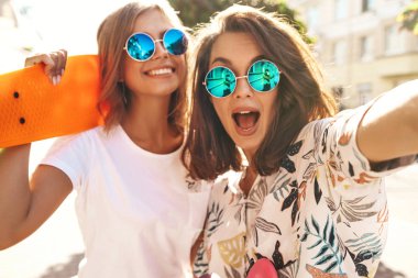 İki genç kadın şık hippi esmer ve sarışın Bayan modelleri yaz güneşli günde selfie sosyal medya için Smartphone cep telefonu ile sokak arka plan üzerinde çekim hipster giysili. Renkli kuruş kaykaylar ile 