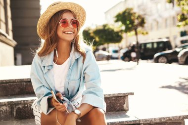 Güzel sevimli gülümseyen sarışın genç model sokak arka merdivenlerde ve müzik dinleme cep telefonu üzerinde oturan yaz hipster giysileri portresi