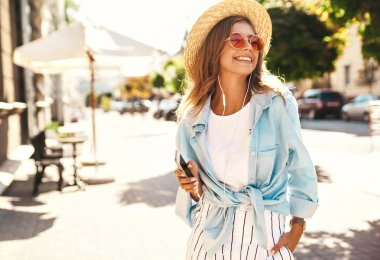 Portre sarışın sevimli gülümseyen güzel, genç model sokak arka plan üzerinde yürüme ve cep telefonu müzik dinleme yaz hipster giysileri