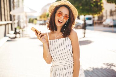 Yaz hipster mutlu gülümseyen genç kız portresi onun kulaklık sokakta smartphone üzerinden müzik dinleme güneşli bir günde giysiler.