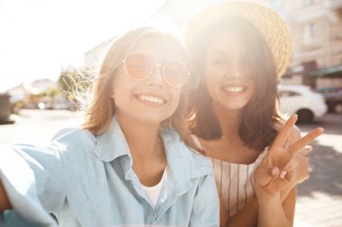 İki genç kadın şık hippi esmer ve sarışın Bayan modelleri yaz güneşli günde selfie sosyal medya için Smartphone cep telefonu ile sokak arka plan üzerinde çekim hipster giysili. Barış işareti gösterilen