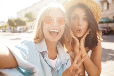 İki genç kadın şık hippi esmer ve sarışın Bayan modelleri yaz güneşli günde selfie sosyal medya için Smartphone cep telefonu ile sokak arka plan üzerinde çekim hipster giysili
