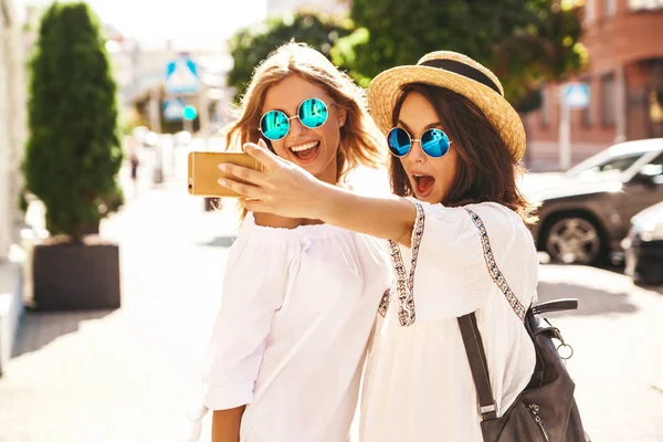 İki genç kadın şık hippi esmer ve sarışın Bayan modelleri yaz güneşli günde selfie sosyal medya için Smartphone cep telefonu ile sokak arka plan üzerinde çekim beyaz hipster giysili