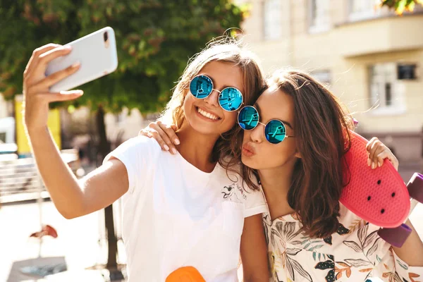 İki genç kadın şık hippi esmer ve sarışın Bayan modelleri yaz hipster giysileri selfie sosyal medya için Smartphone cep telefonu ile sokak arka plan üzerinde çekim. Renkli kuruş kaykaylar ile 