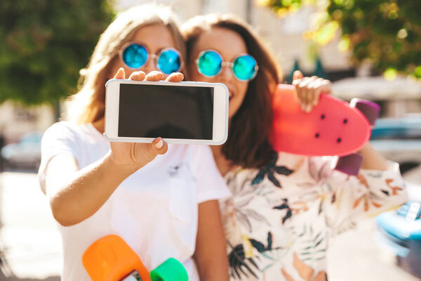 Две молодые женщины стильные брюнетки хиппи и блондинки модели летом солнечный день в хипстерской одежде делает селфи фотографии для социальных сетей на смартфоне на улице. С разноцветными скейтбордами
 