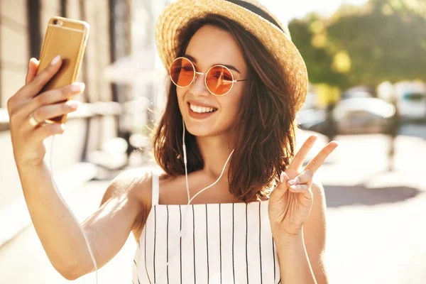 青年微笑的女孩在夏天白色时髦的衣裳在耳机采取自拍在温暖的日落背景智能手机上 — 图库照片