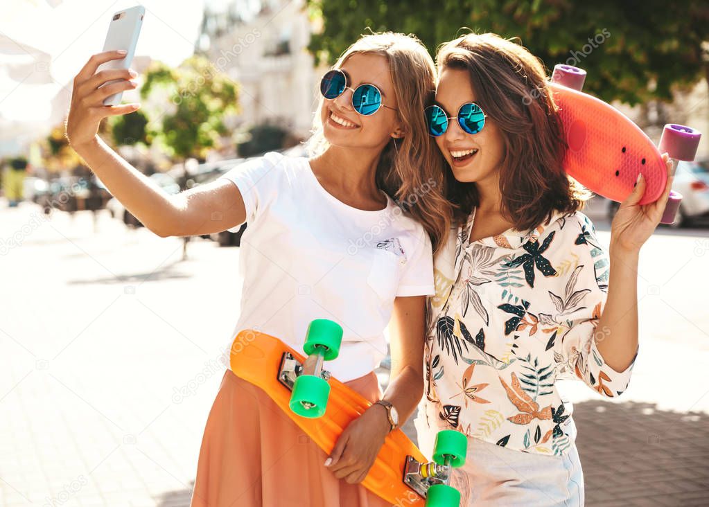 Two young female stylish hippie brunette and blond women models in summer sunny day in hipster clothes taking selfie photos for social media on smartphone on the street background. With colorful penny skateboards 