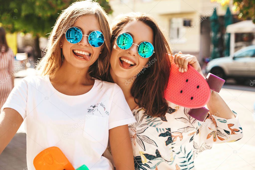 Two young female stylish hippie brunette and blond women models in summer hipster clothes taking selfie photos for social media on smartphone on the street background. With colorful penny skateboards. Show tongue