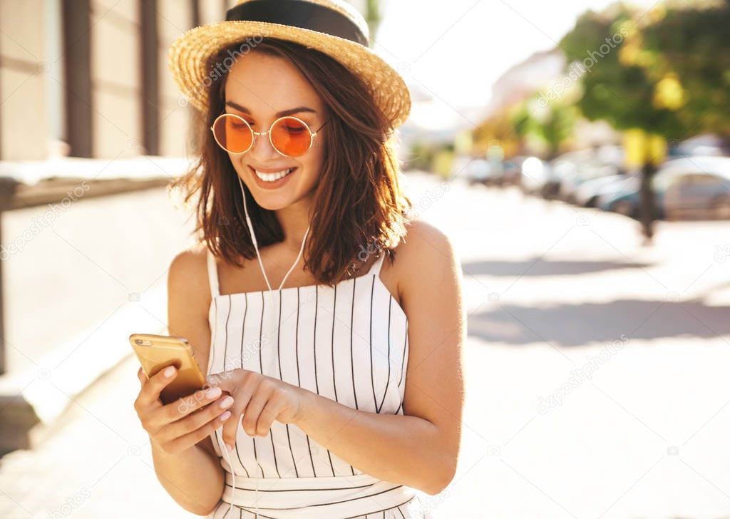 Portrait of happy smiling teen girl in summer hipster clothes listening music on her headphones from smartphone in the street in a summer sunny day