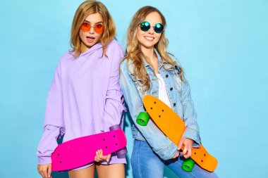 İki genç sarışın kadınlarla kuruş kaykaylar gülümseyen şık. Modelleri yaz hipster spor giysileri Studio mavi duvarının yakınında poz. Pozitif kızlar çıldırıyor