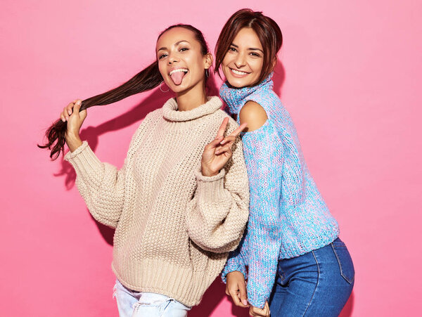 Two beautiful sexy smiling gorgeous girls looking at camera. Hot women standing in stylish white and blue sweaters, on pink background. Showing peace sign