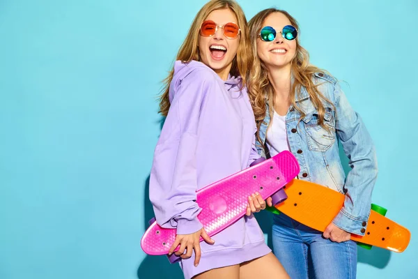 二人の若いペニー スケート ボードで金髪の女性の笑顔がスタイリッシュ スタジオで青い壁の近くポーズ夏流行に敏感なスポーツ服の女の子 肯定的なモデル — ストック写真