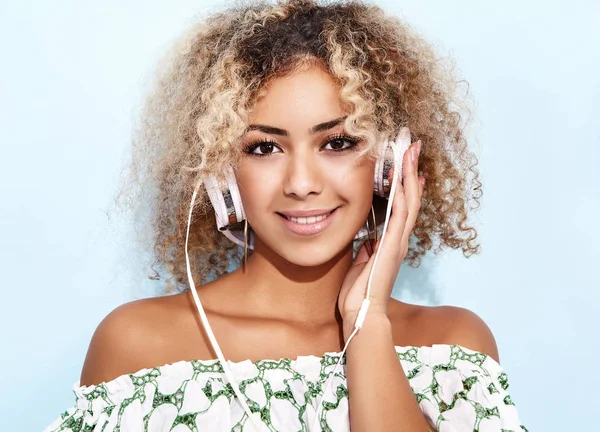 Fashion happy beautiful black woman with afro blond hair.Girl smiling and listening to music in headphones. Playful hipster model posing near blue wall