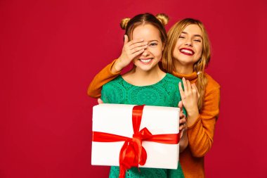 Noel, x-mas, kavram. İki şık giysiler güzel kadın gülümsüyor. Kızlar kırmızı zemin üzerine poz. Kaplama gözleri onun arkadaşı ve onun büyük hediye kutusu veren model. Eğleniyor, kutlama için hazır.
