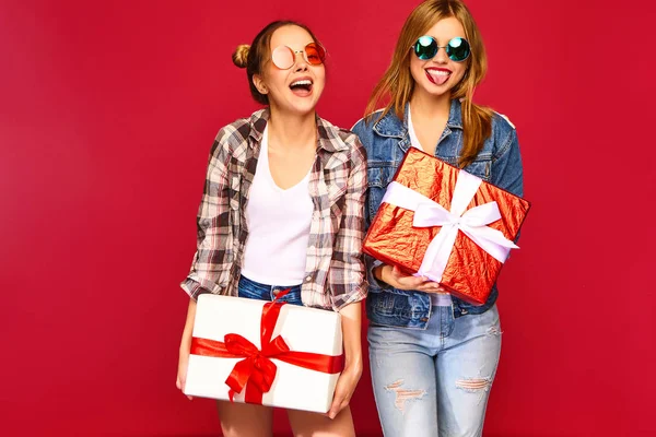 圣诞节 两个穿着时尚服装的微笑的美女 女孩在红色背景摆姿势 带有大礼品盒的模型 玩得开心 准备庆祝 最好的朋友的明亮假期 — 图库照片