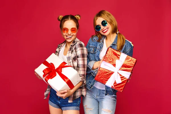 圣诞节 两个穿着时尚服装的微笑的美女 女孩在红色背景摆姿势 带有大礼品盒的模型 玩得开心 准备庆祝 最好的朋友的明亮假期 — 图库照片
