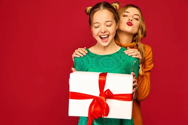 圣诞节 两个穿着时尚服装的微笑的美女 女孩在红色背景摆姿势 模特给她的朋友大礼物盒 玩得开心 准备庆祝 — 图库照片