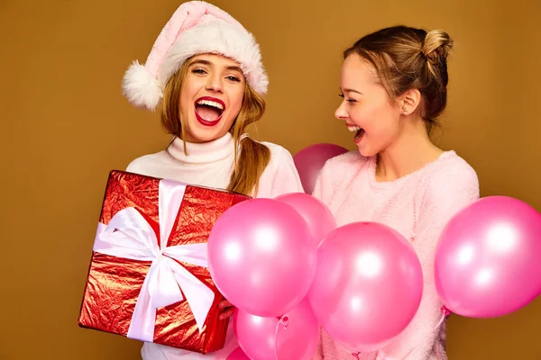 圣诞节 两个面带微笑的美丽女人穿着白色的冬衣 女孩在金色背景上摆姿势 带有大礼品盒和粉红色气球的模型 玩得开心 准备庆祝新年 — 图库照片